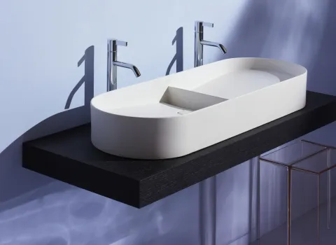 Ein modernes Badezimmer zeigt ein elegantes, ovales Waschbecken mit zwei verchromten Wasserhähnen an einer schwarzen, schwebenden Ablage.