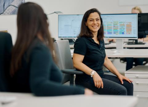 Eine Frau sitzt lächelnd an einem Schreibtisch in einem modernen Büro und unterhält sich mit einer Kollegin, die im Vordergrund unscharf zu sehen ist. 