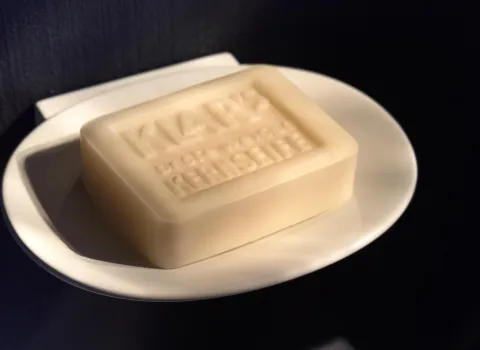 Ein rechteckiges Stück helle Seife liegt auf einer weißen Seifenschale, deren glatte Oberfläche das Licht reflektiert. Auf der Seife ist der Schriftzug "Klar's" und "Reine Pflanzenölkernseife" eingeprägt.