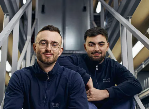 Zwei junge Männer in Arbeitskleidung von bad und heizung sitzen auf einer Metalltreppe in einem Lagerraum, umgeben von Regalen voller Ersatzteile und Verpackungen. 